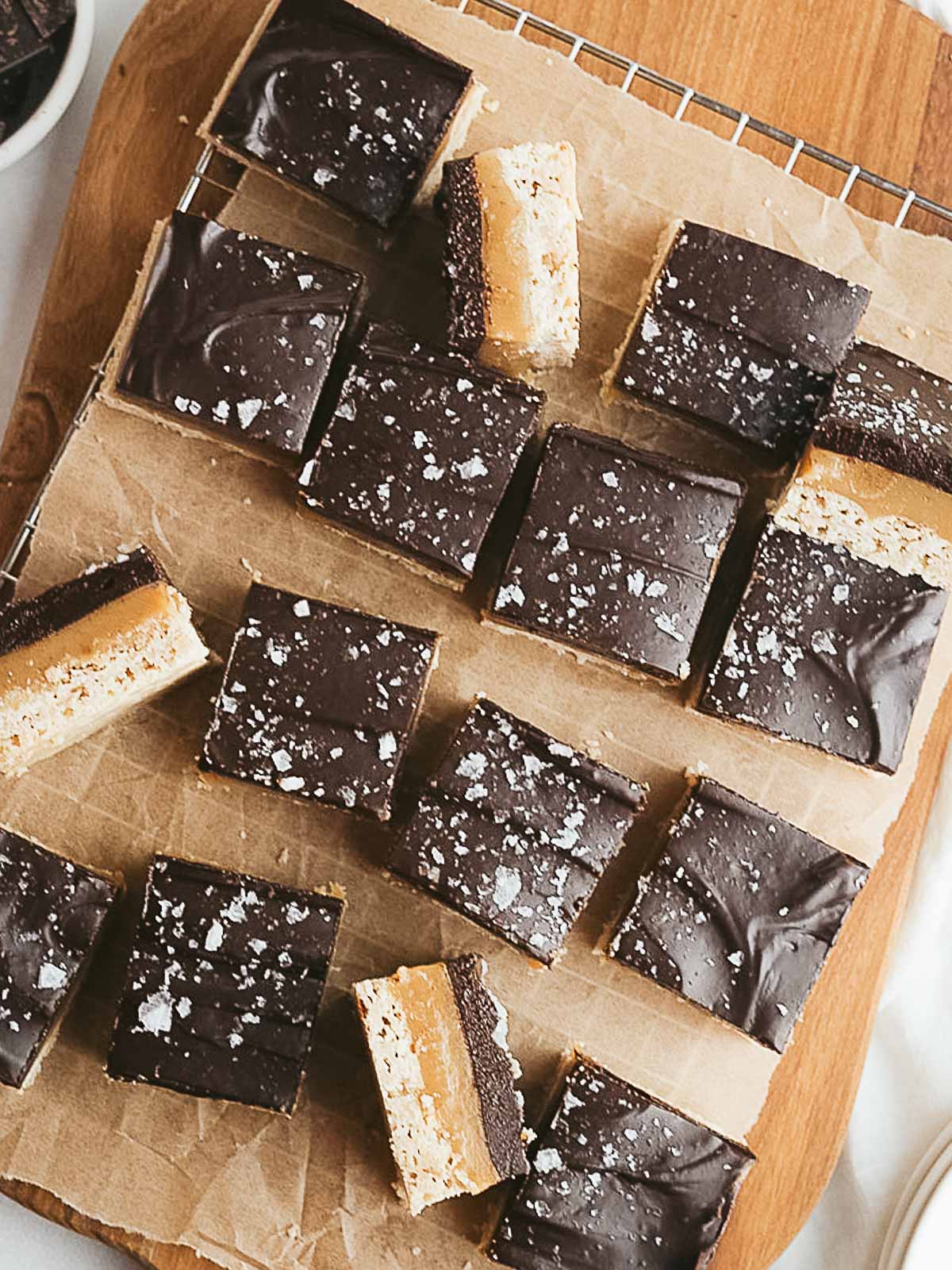 Squares of chocolate and caramel layered bars topped with flaky sea salt are arranged on a parchment-lined wooden board.