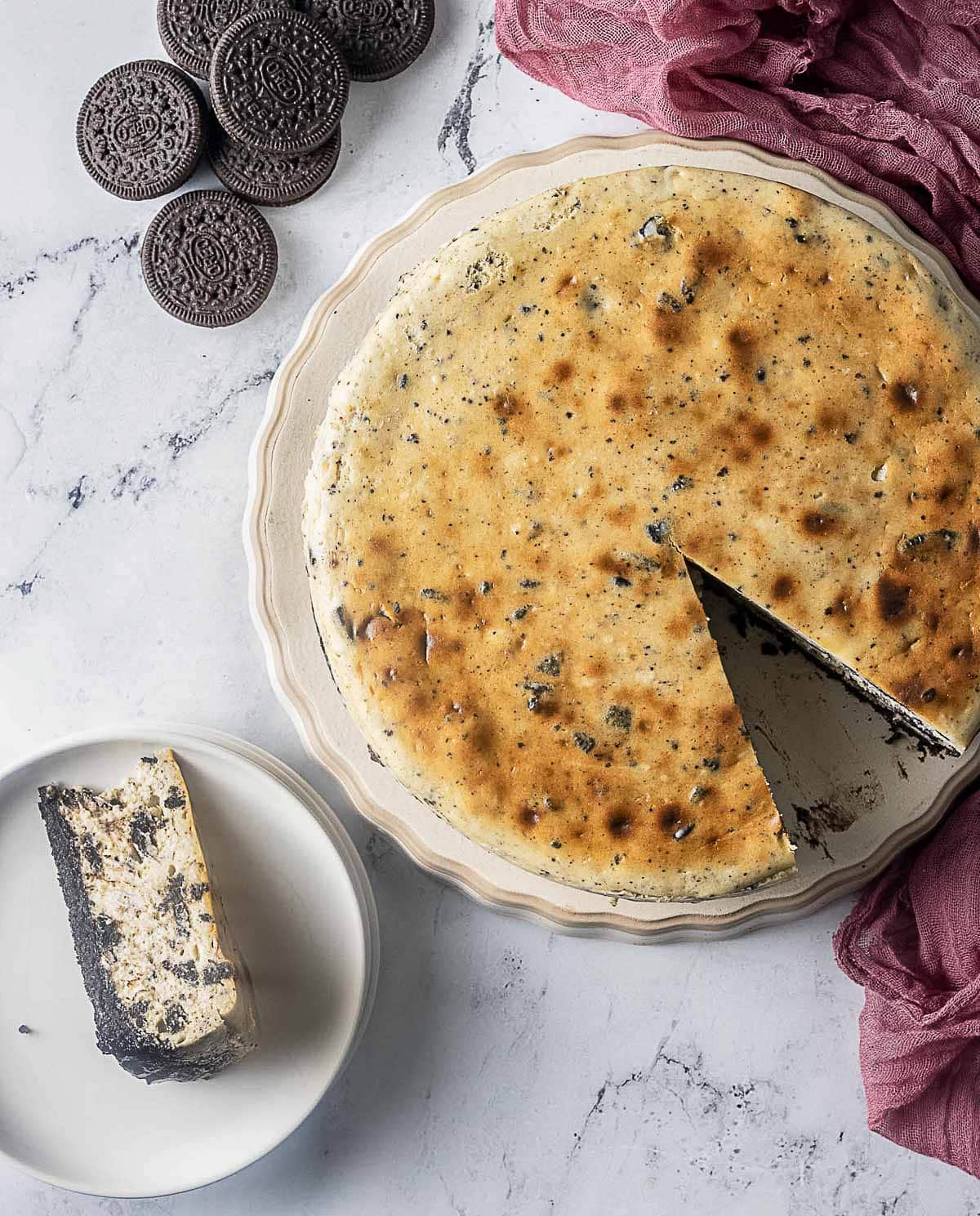 A baked Oreo cheesecake with one slice cut out, served on a white plate. Whole Oreo cookies are scattered nearby on a marble surface.