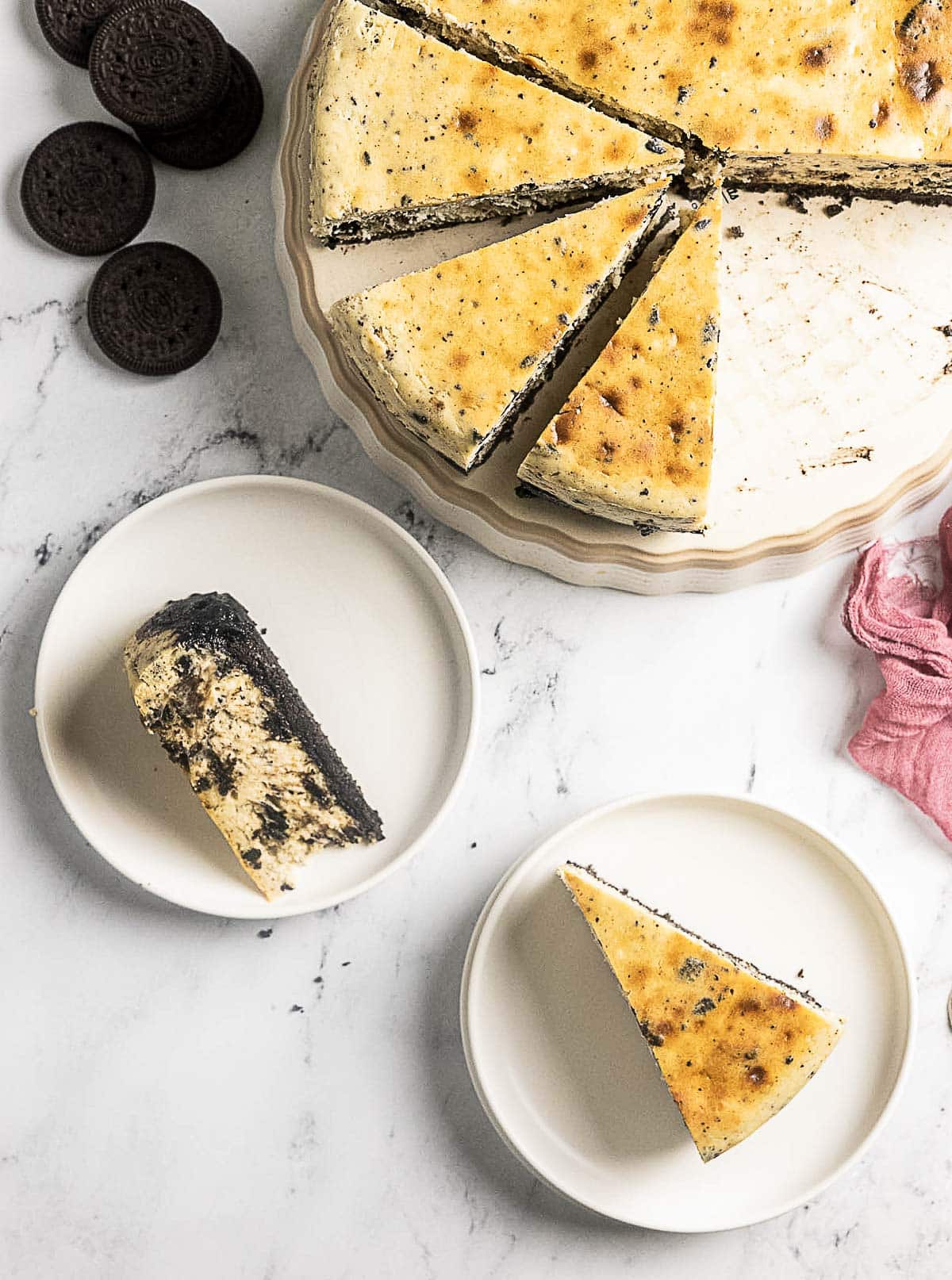 A sliced cookies and cream cheesecake on a cake stand, with two slices served on plates and several chocolate sandwich cookies nearby on a marble surface.