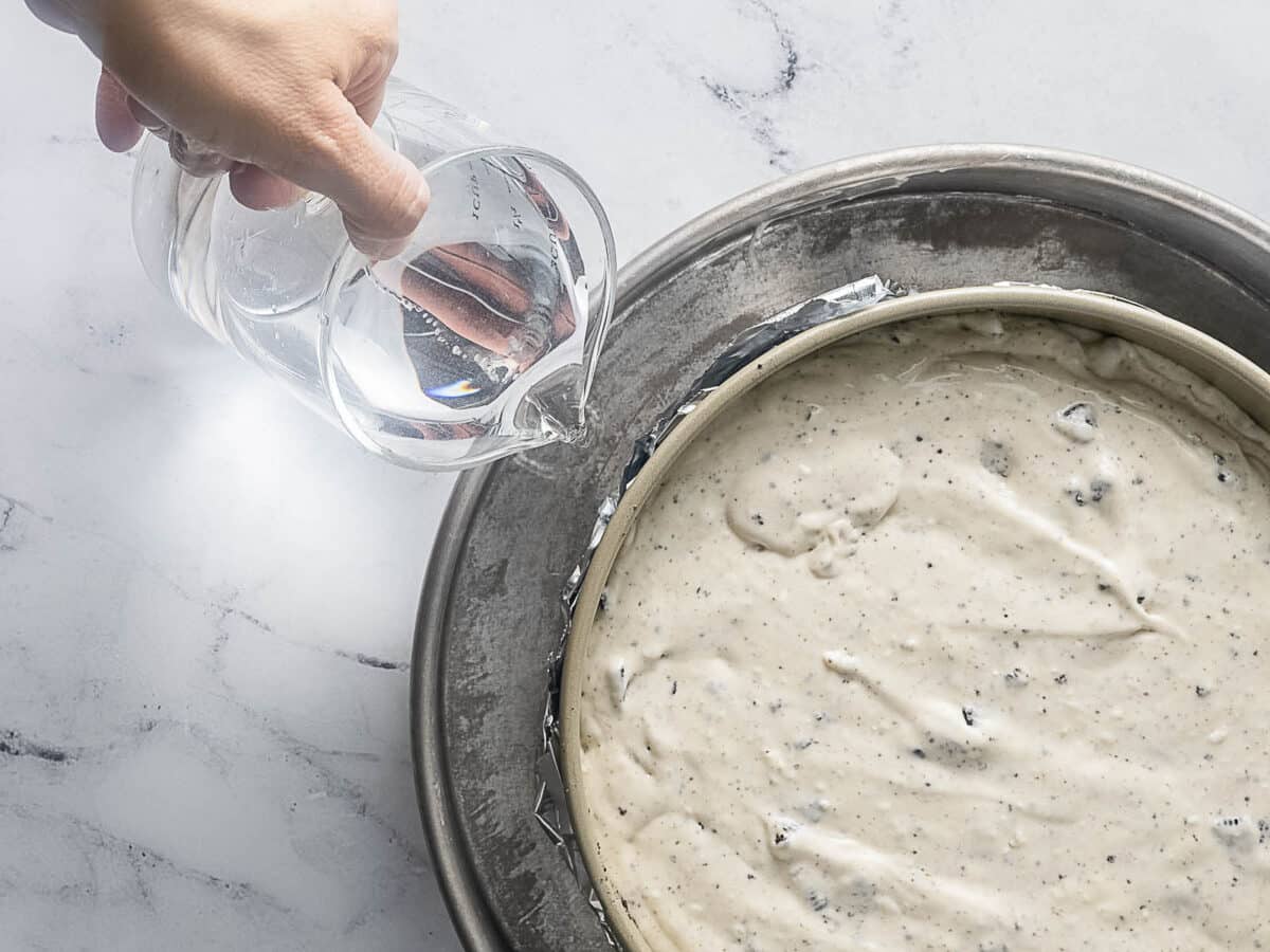 A hand pours water from a measuring cup into a pan surrounding a springform pan filled with cake batter.