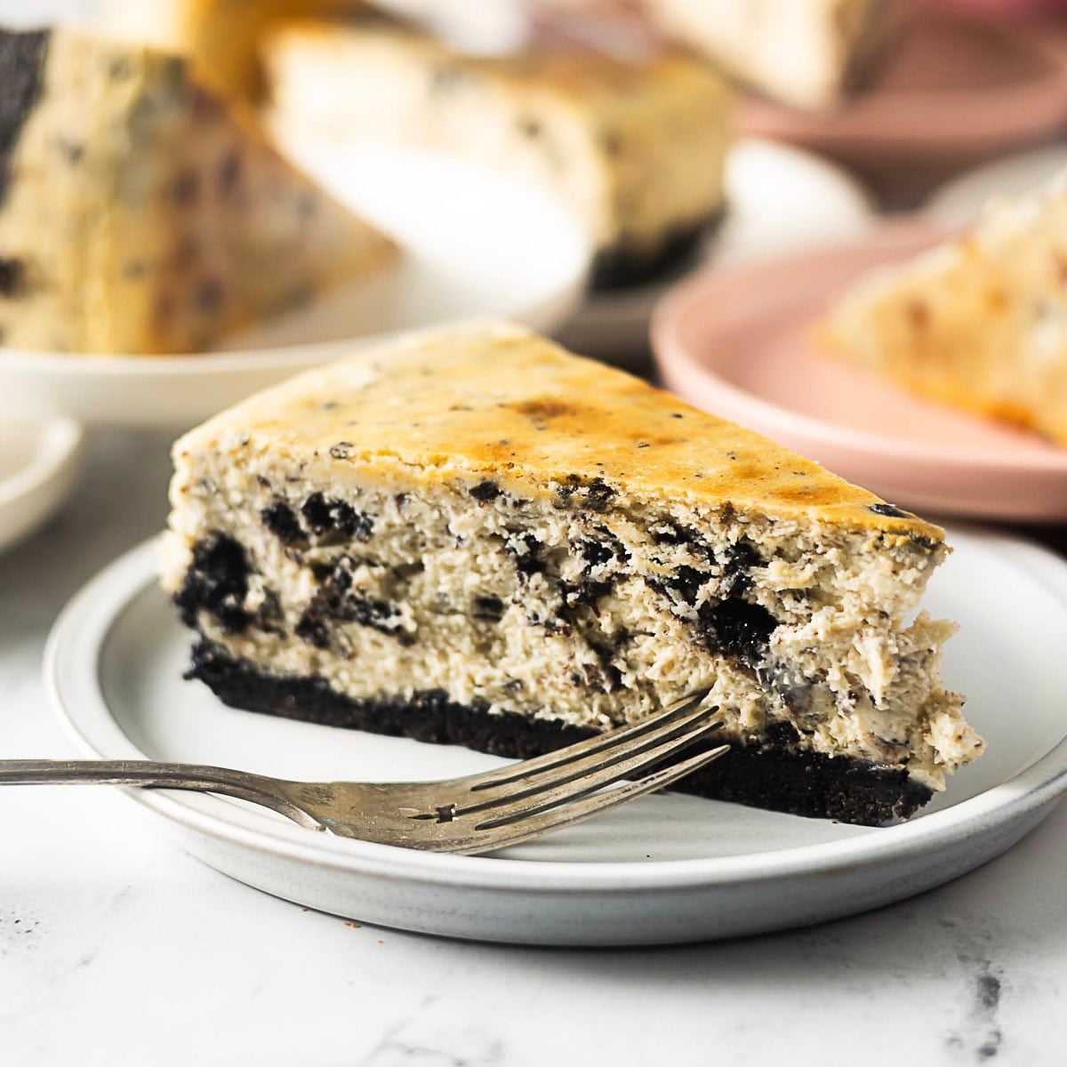 A slice of cookies and cream cheesecake with a dark cookie crust sits on a white plate with a fork.