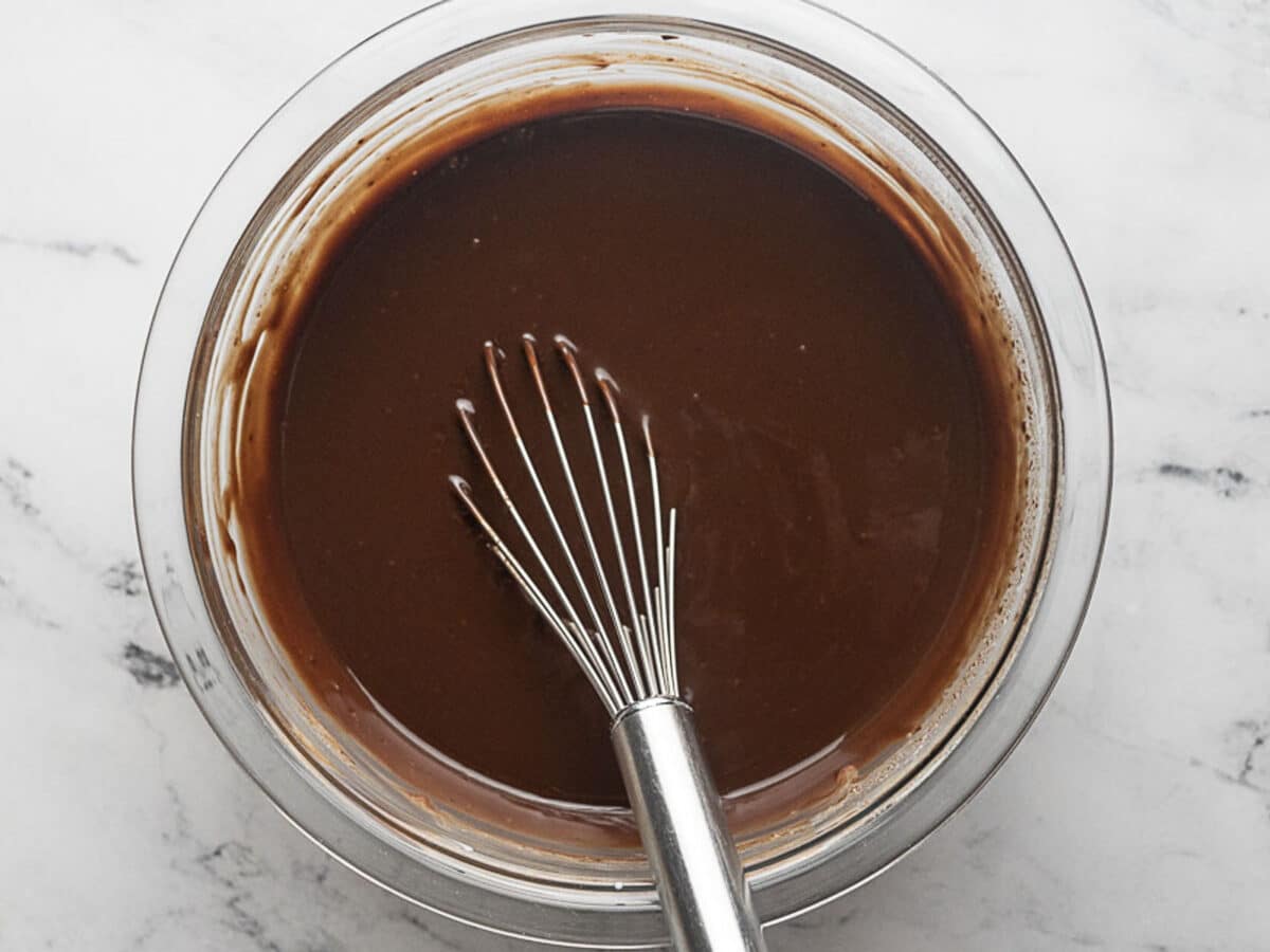 A metal whisk rests in a glass bowl filled with smooth chocolate batter on a white marble surface.