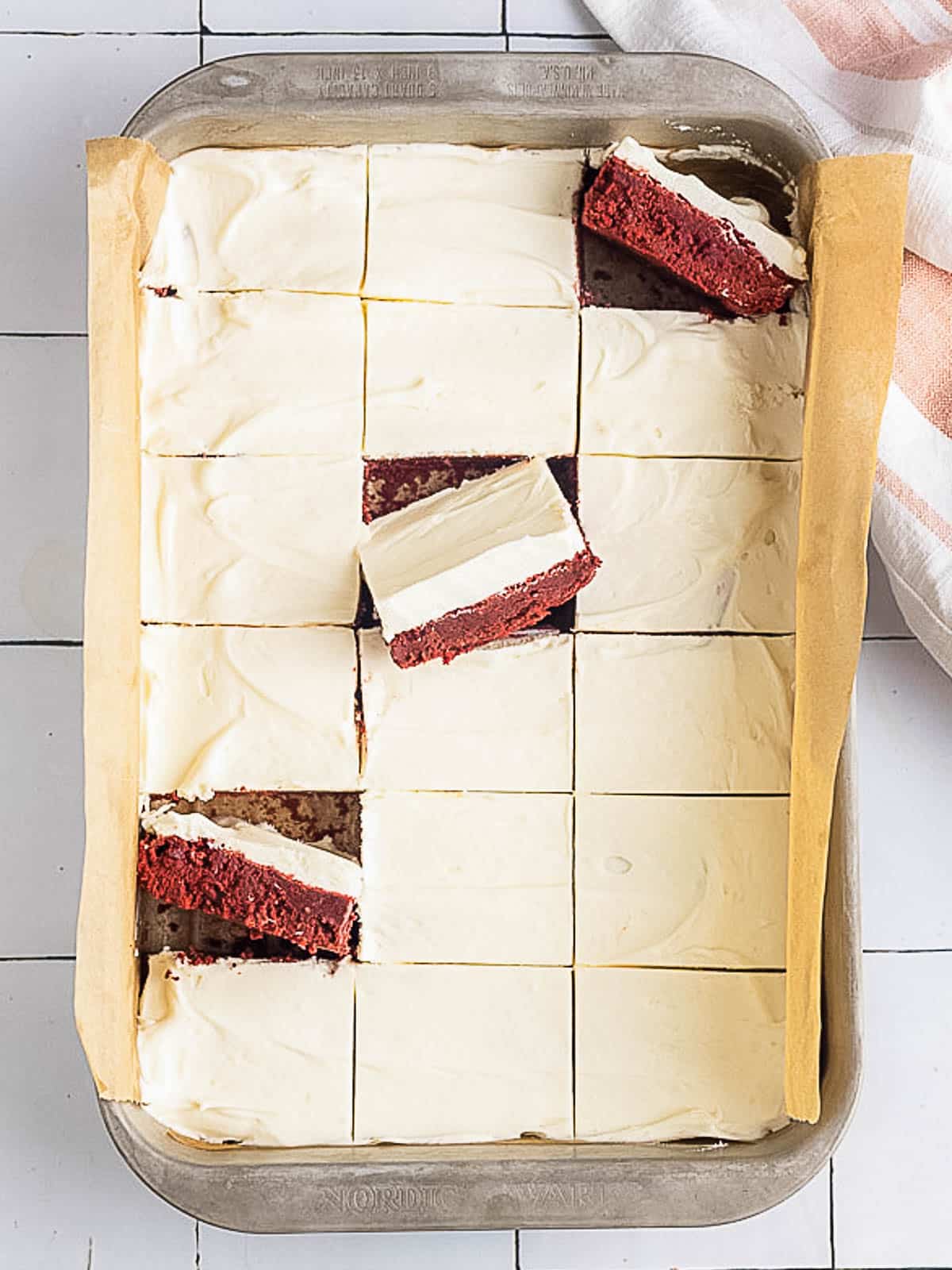A pan of 15 square red velvet cake bars topped with cream cheese frosting, some pieces slightly tilted to show the red cake layer beneath.