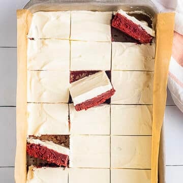 Tray of red velvet cheesecake bars, cut into squares, with parchment lining the pan; three bars are stacked on top of each other, showing the red and cream layers.