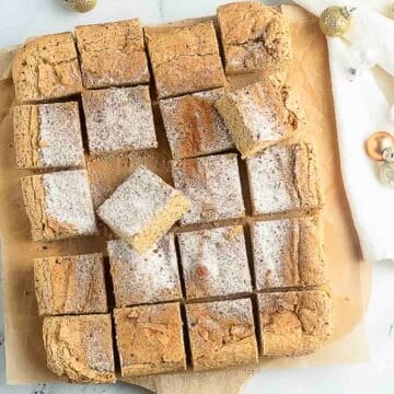 A batch of sixteen sugar-dusted, square cinnamon sugar snickerdoodles bars arranged on parchment paper, with some holiday decorations nearby.