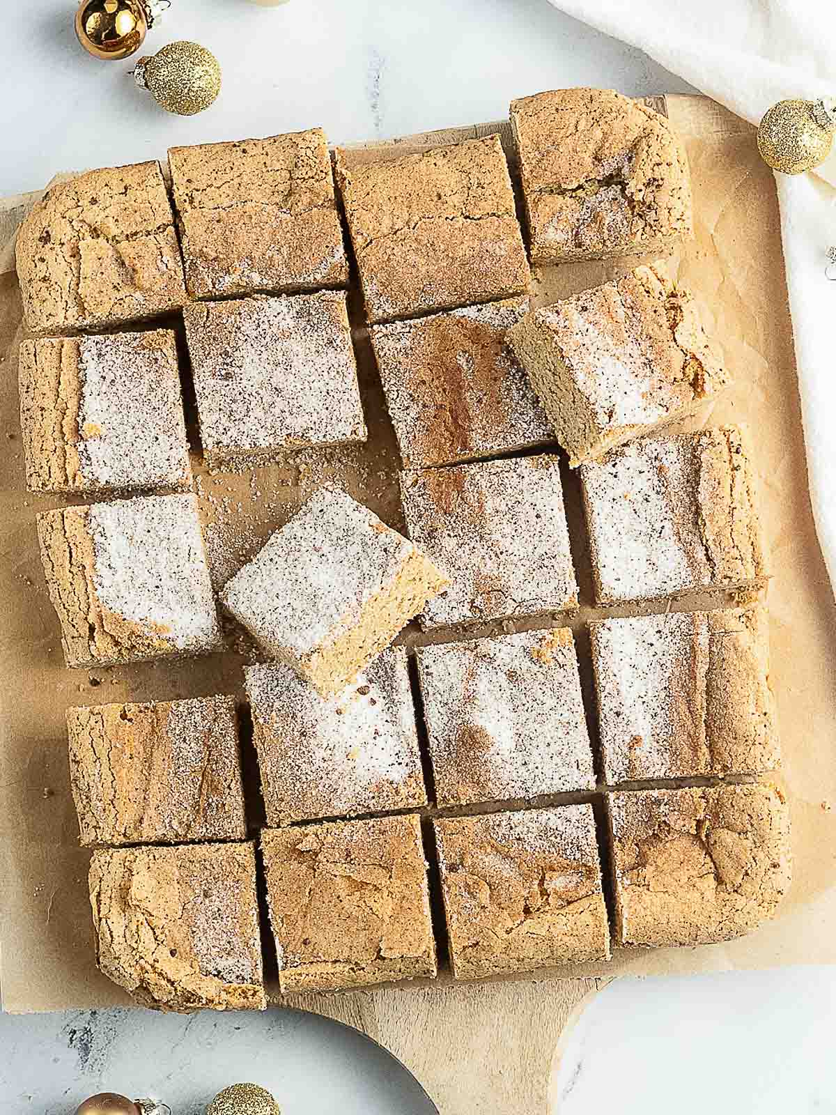 A batch of snickerdoodle bars cut into squares on parchment paper, some dusted with powdered sugar and cinnamon, with festive gold decorations nearby.