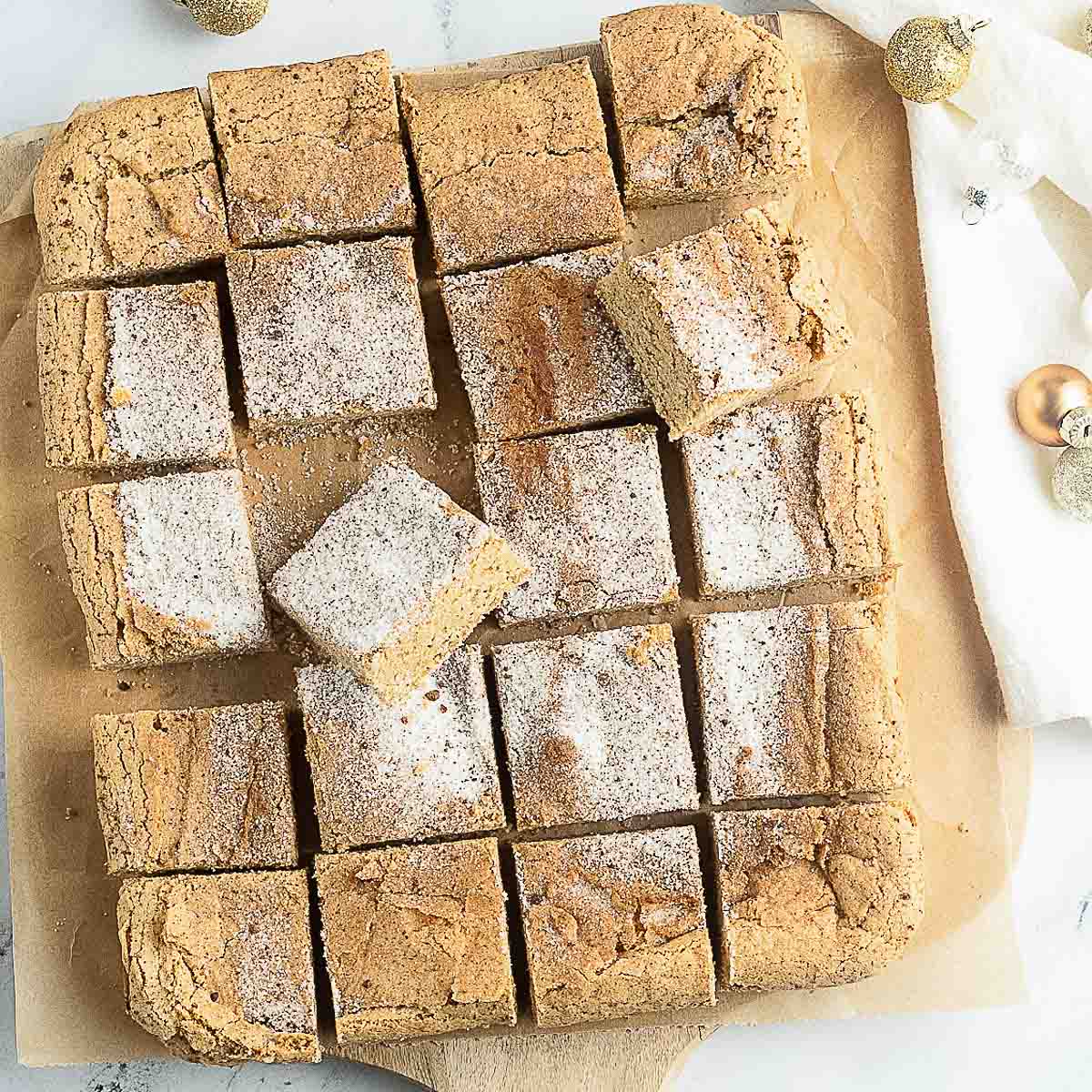 A batch of sixteen sugar-dusted, square cinnamon sugar snickerdoodles bars arranged on parchment paper, with some holiday decorations nearby.