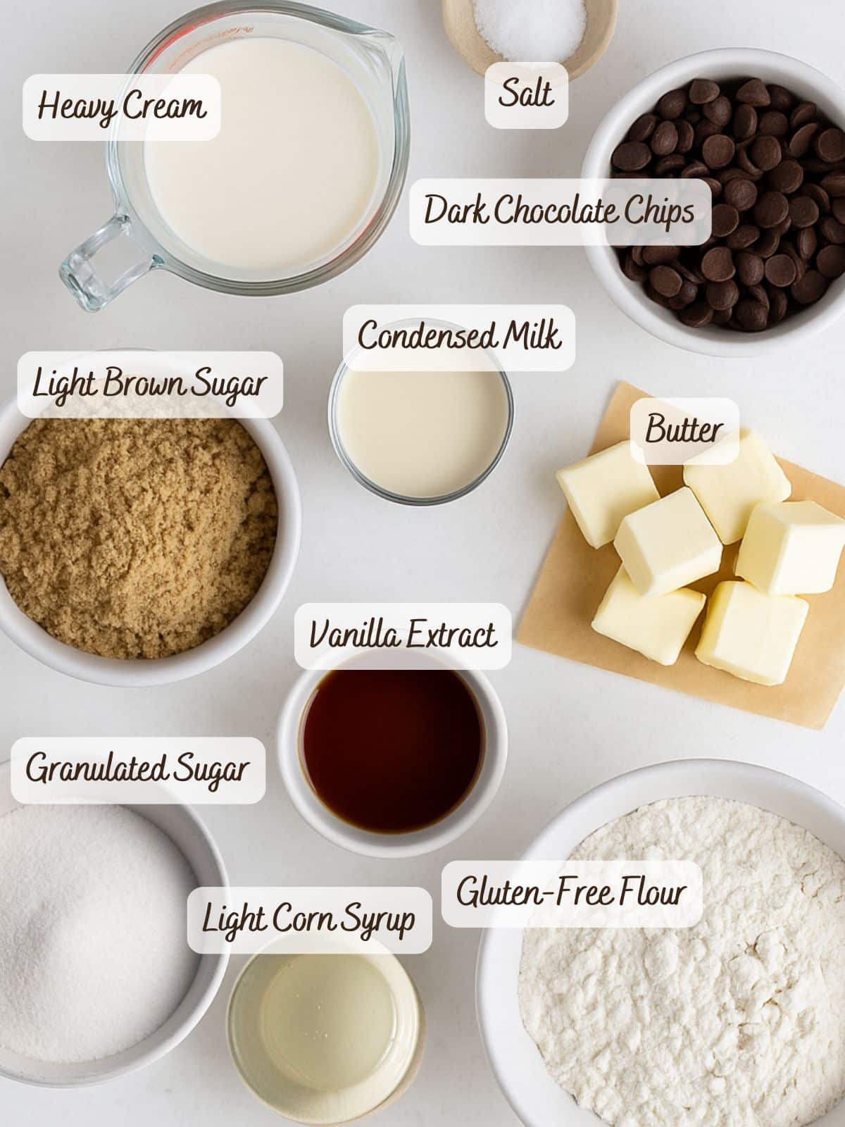 Assorted baking ingredients including heavy cream, salt, dark chocolate chips, two types of sugar, condensed milk, butter, vanilla extract, corn syrup, and gluten-free flour-perfect for making decadent gluten free millionaire shortbread.