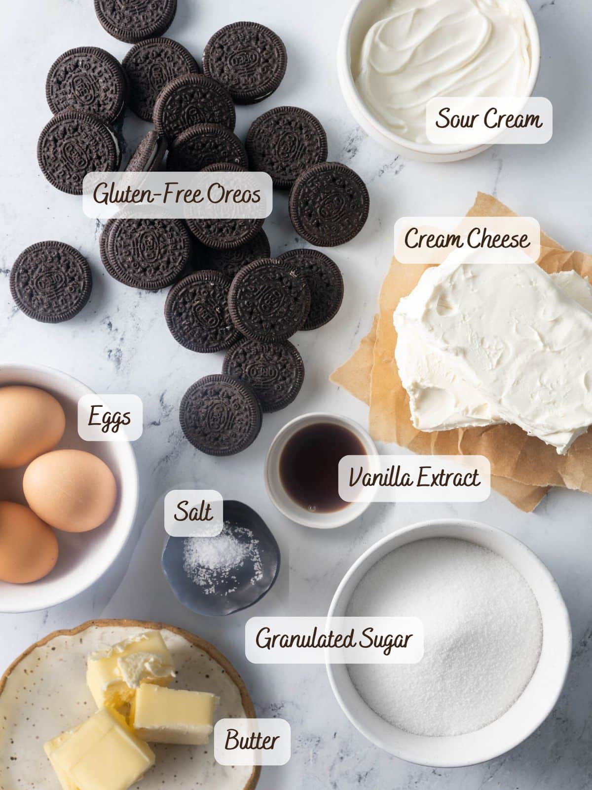 Top-down view of baking ingredients for Gluten Free Oreo Cheesecake, including eggs, gluten-free Oreos, sour cream, cream cheese, vanilla extract, salt, granulated sugar, and butter-all clearly labeled.