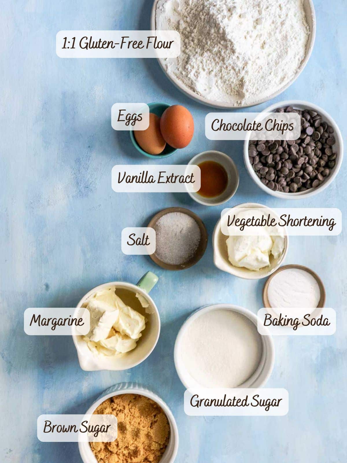 Top-down view of labeled baking ingredients for gluten free dairy free chocolate chip cookies in bowls and cups on a blue surface, including gluten-free flour, chocolate chips, eggs, vanilla extract, shortening, margarine, sugars, salt, and baking soda.