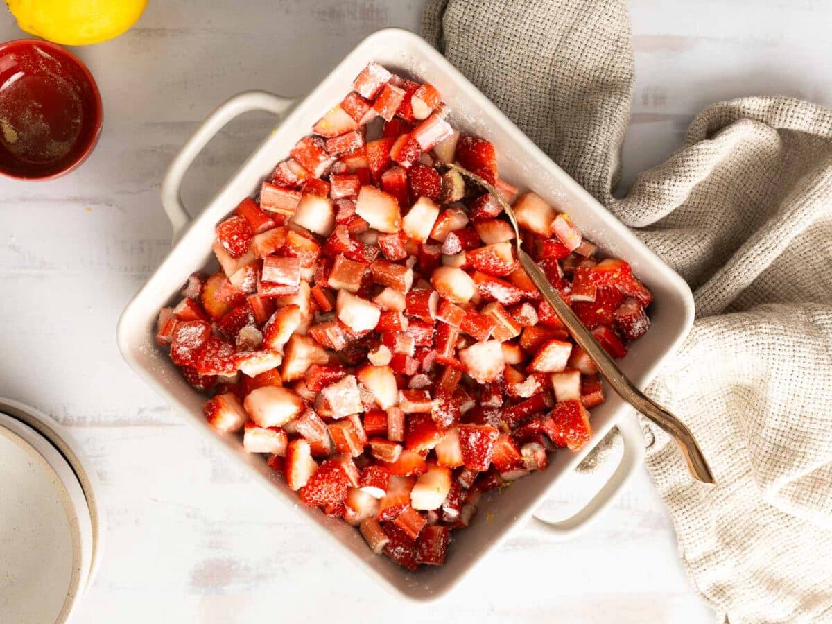 A square white baking dish filled with chopped strawberries and sugar, with a metal spoon and a textured beige cloth nearby on a white surface.