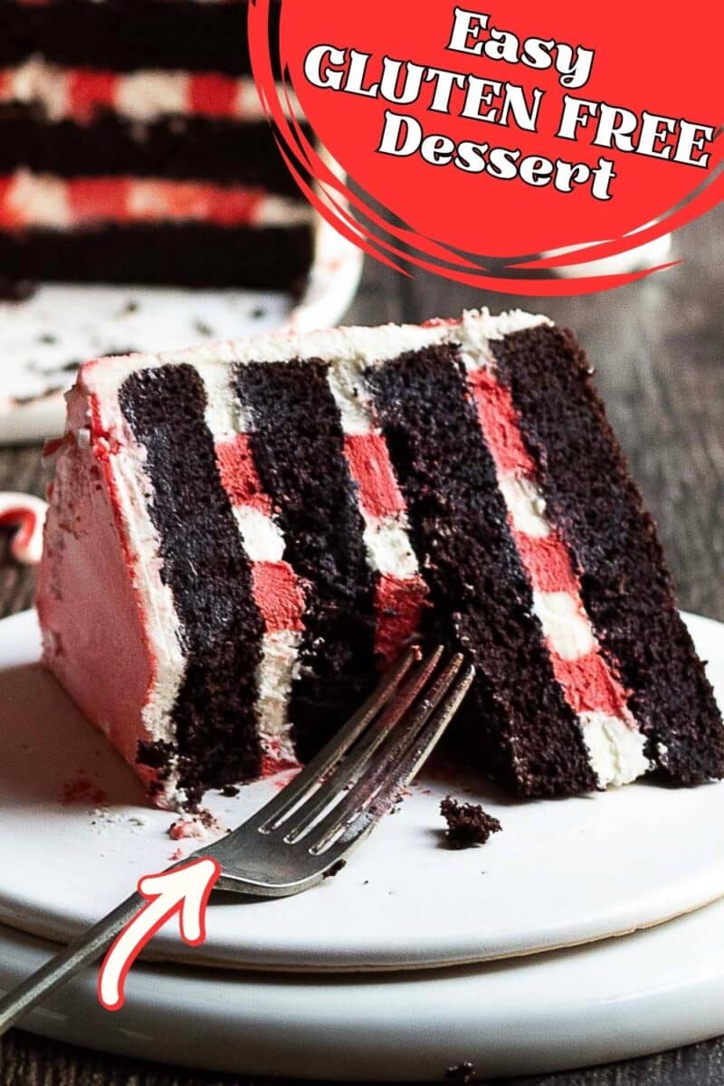 A slice of chocolate and pink layered gluten free peppermint cake with frosting sits on a white plate with a fork
