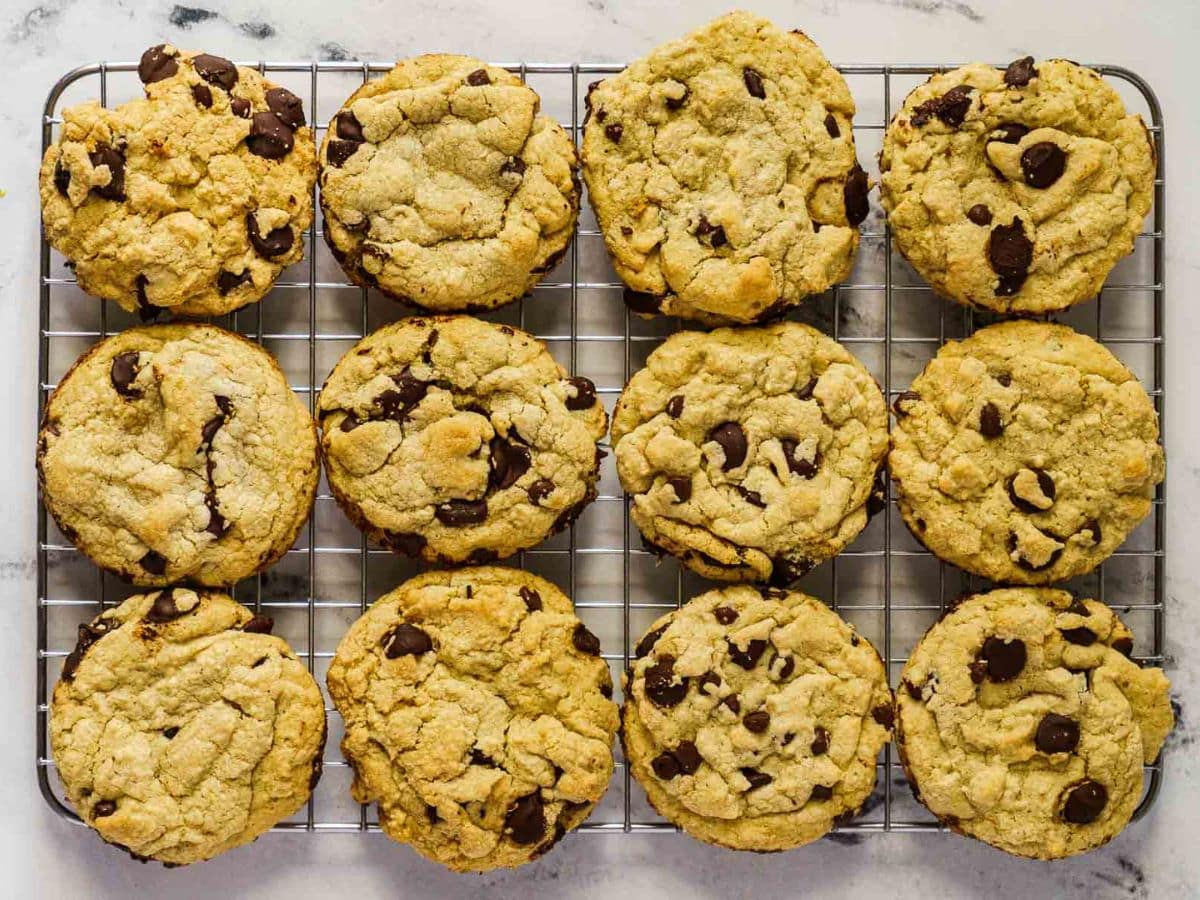 Twelve chocolate chip cookies are arranged in rows on a metal cooling rack placed on a white surface.