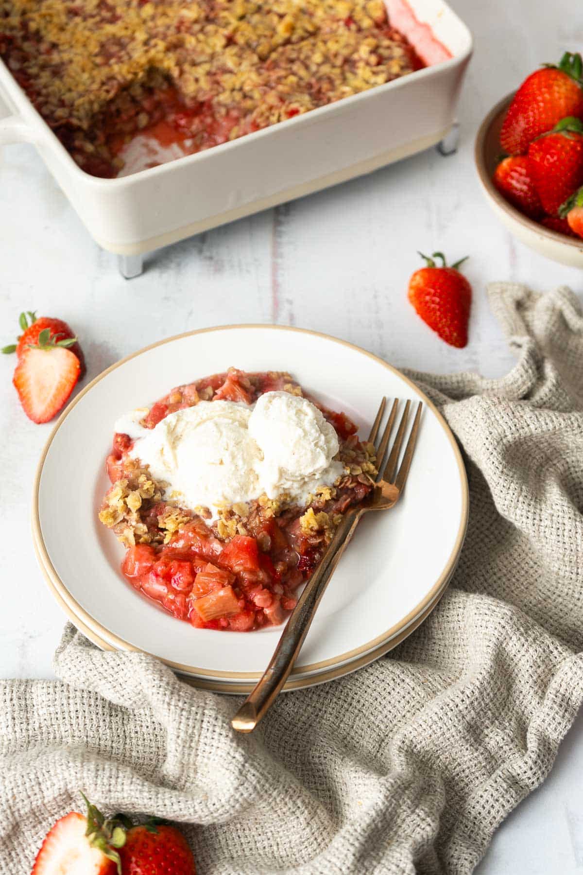 A plate with strawberry rhubarb crisp topped with two scoops of vanilla ice cream sits next to a fork; a baking dish and fresh strawberries are in the background.