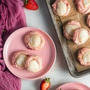 Pink cookies with white icing are arranged on a pink plate and a baking sheet lined with parchment paper; strawberries and a pink cloth are nearby.