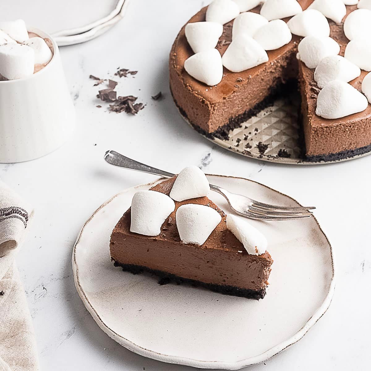 A slice of chocolate cheesecake topped with marshmallows sits on a plate with a fork, next to the rest of the cheesecake and a cup of dessert in the background.