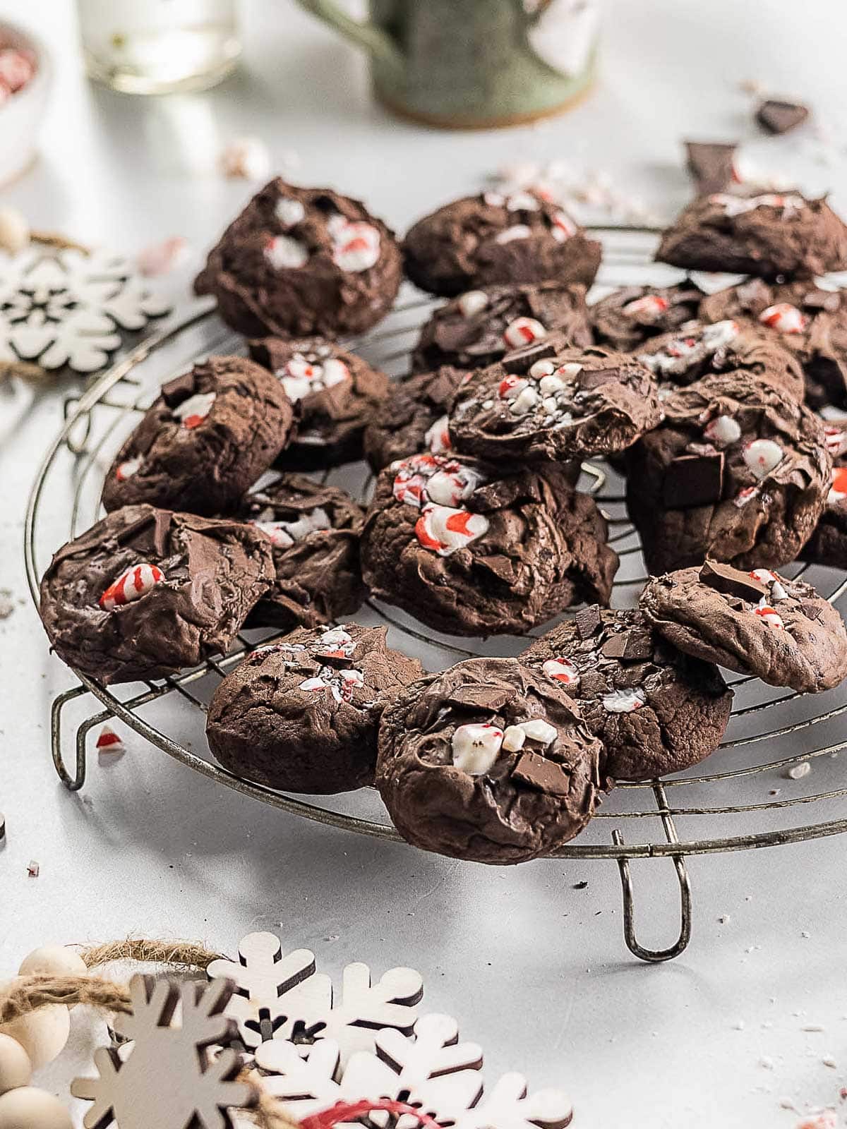 A cooling rack holds several chocolate cookies topped with crushed peppermint candies, with festive decorations nearby.