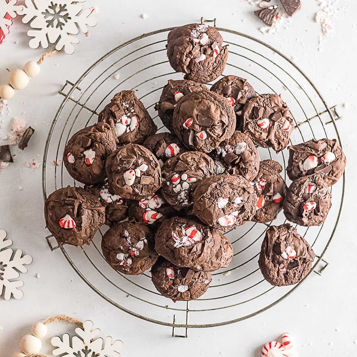 A wire cooling rack holds a pile of chocolate cookies topped with crushed peppermint candies, surrounded by snowflake decorations and beads on a white surface.