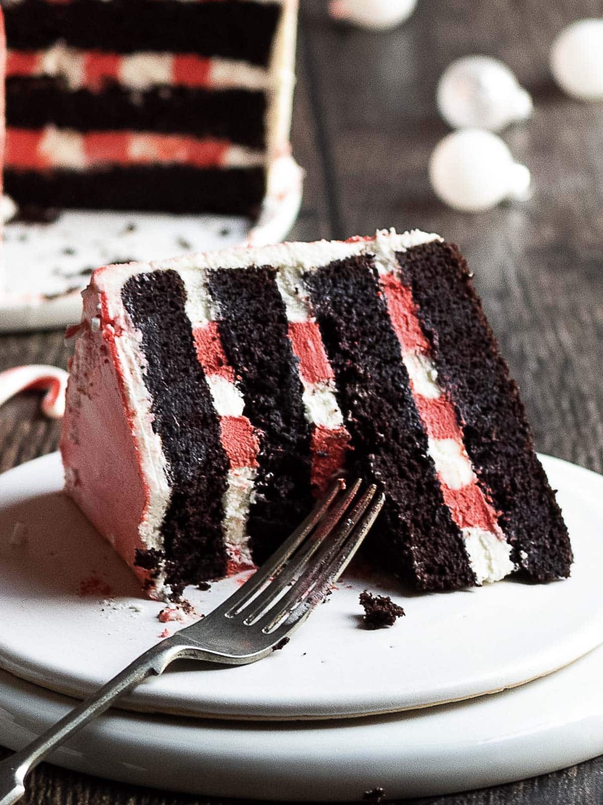 A slice of layered chocolate cake with red and white filling sits on a plate with a fork, with more cake visible in the background.