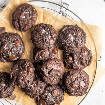 A batch of chocolate cookies with chocolate chips and sea salt on top, arranged on parchment paper over a cooling rack.