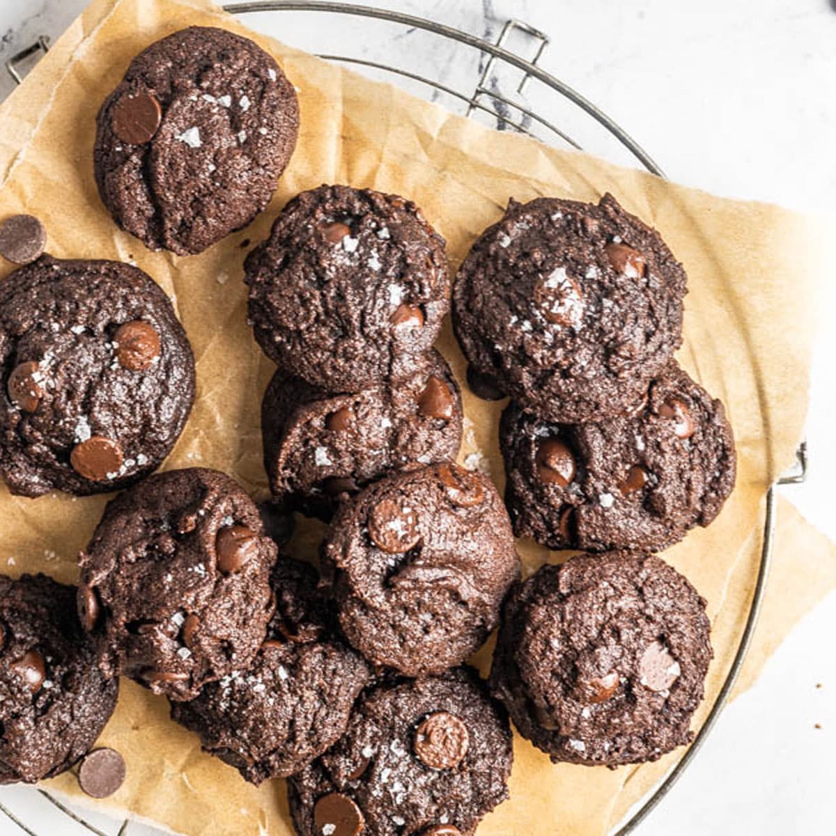 A batch of chocolate cookies with chocolate chips and sea salt on top, arranged on parchment paper over a cooling rack.