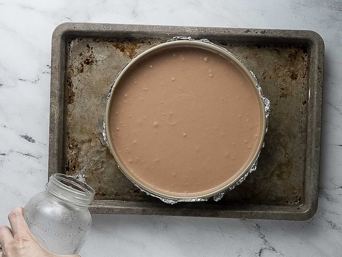 A round cake pan filled with chocolate batter sits on a metal baking sheet, with a hand pouring water from a glass jar into the tray.