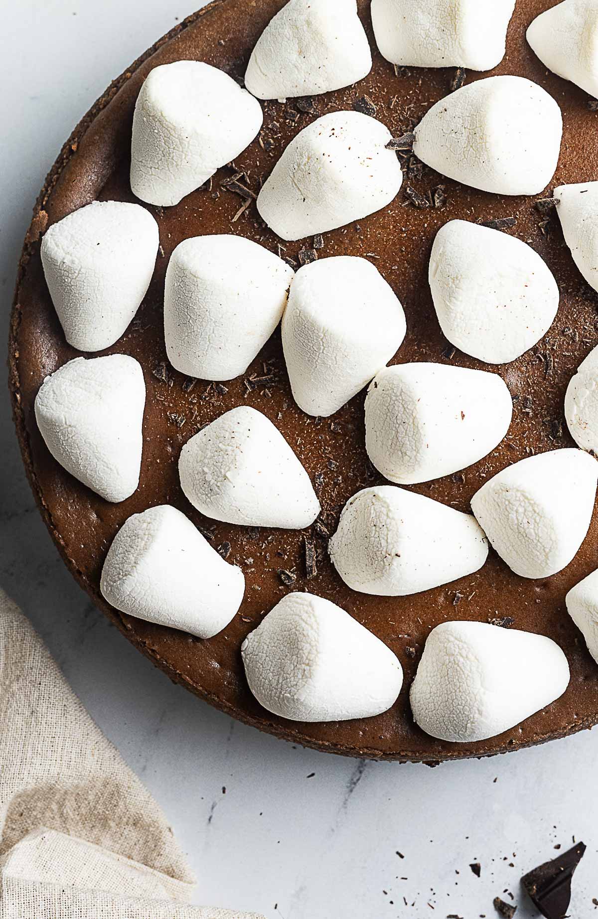 A chocolate pie topped with large white marshmallows, shown partially from above on a light surface with a beige napkin nearby.