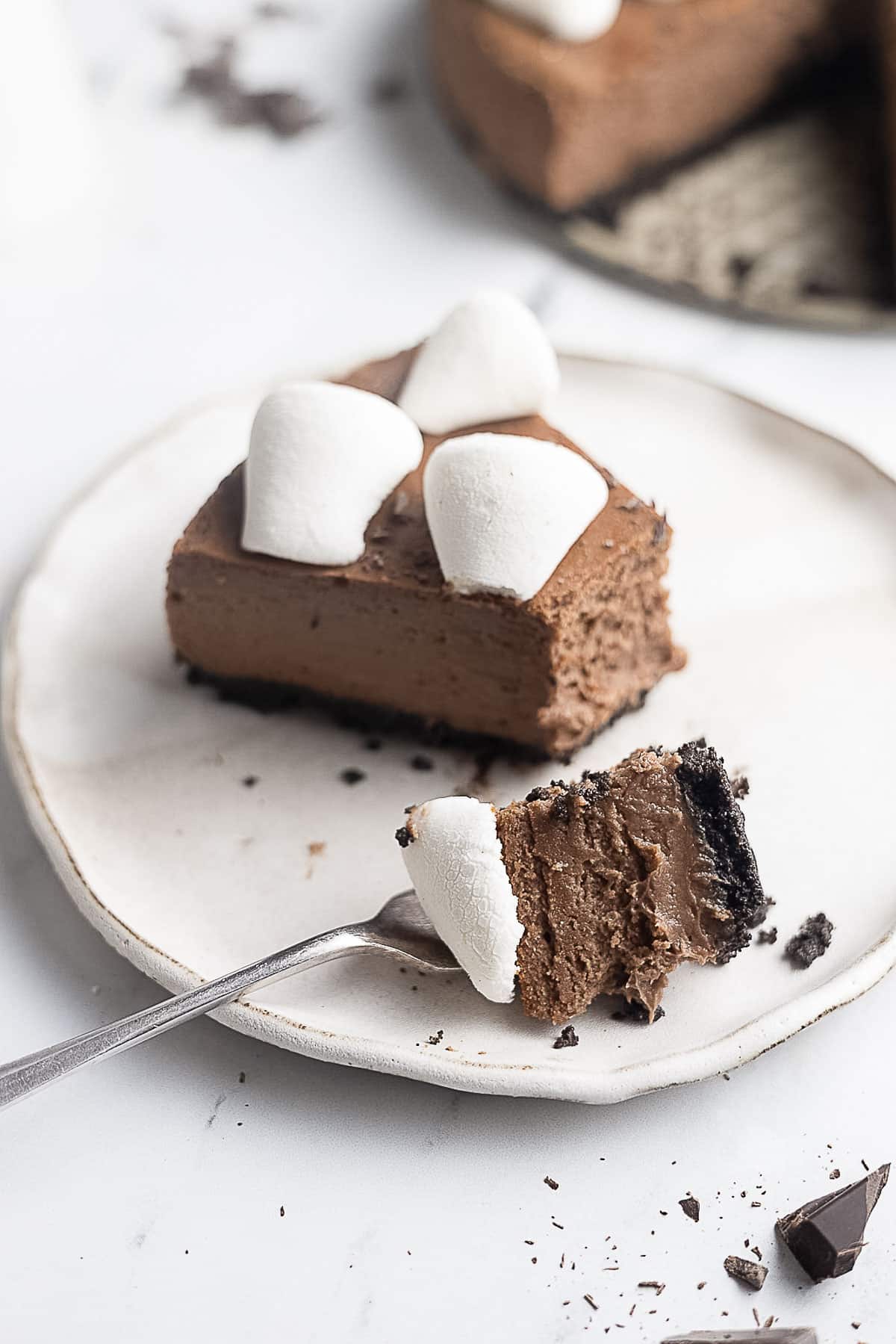 A slice of chocolate cheesecake with an Oreo crust and marshmallows on top, served on a white plate, with a fork holding a bite-sized piece.