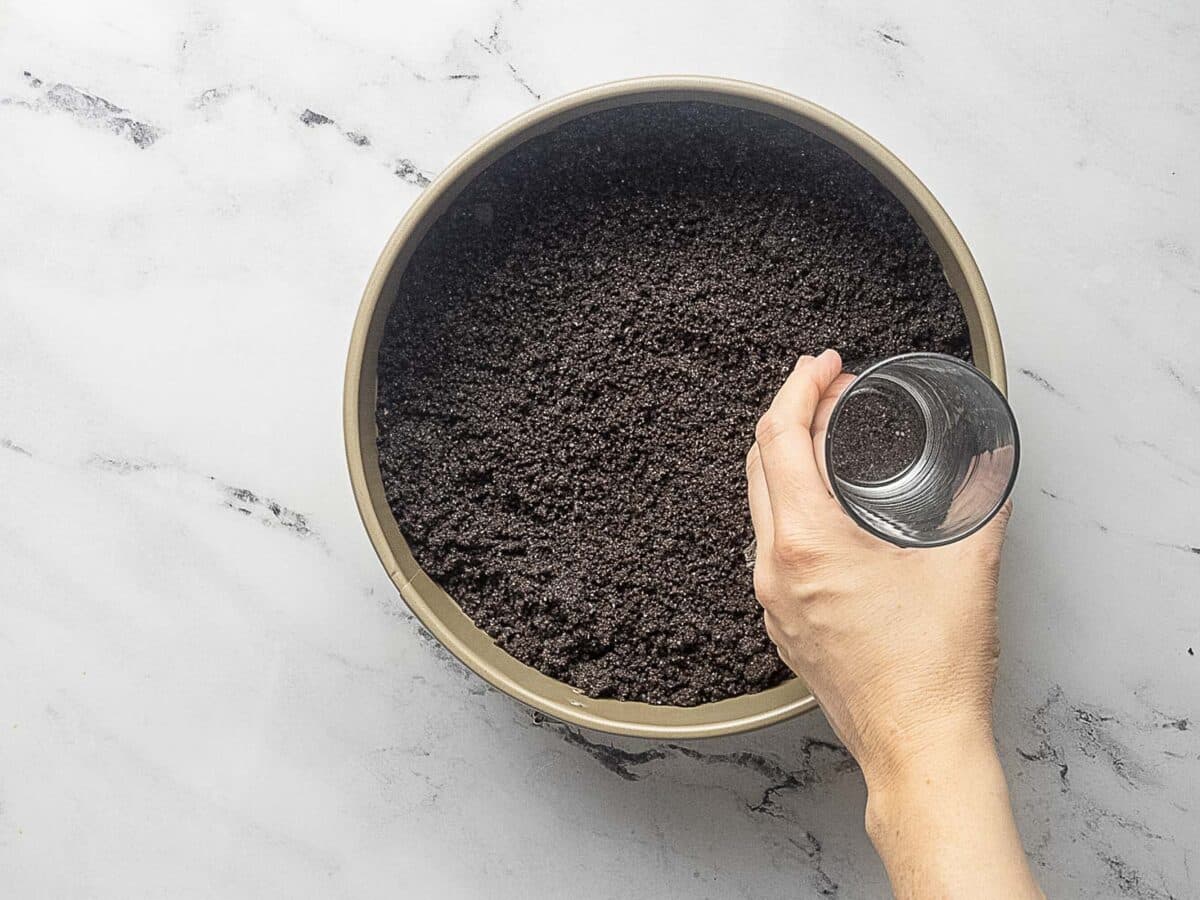 A hand presses a glass into a pie pan filled with crushed chocolate cookies to form a crust on a marble countertop.