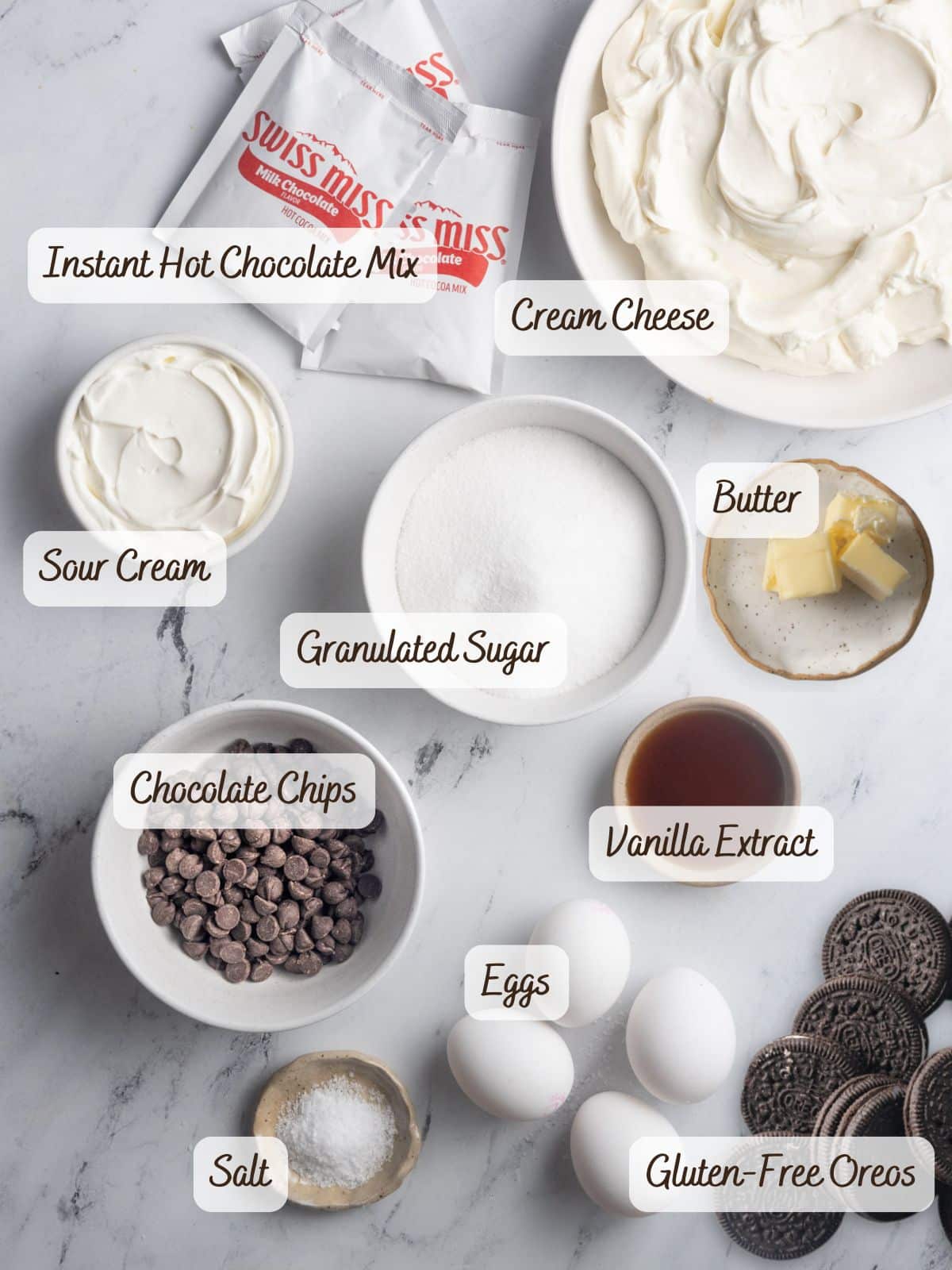 A flat lay of baking ingredients for a gluten free chocolate cheesecake, labeled: instant hot chocolate mix, cream cheese, sour cream, granulated sugar, butter, chocolate chips, vanilla extract, eggs, salt, and gluten-free Oreos.