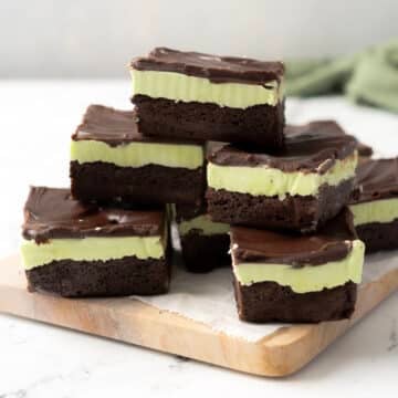 Several layered mint chocolate brownies with a green mint filling and chocolate topping are stacked on a wooden board lined with parchment paper.