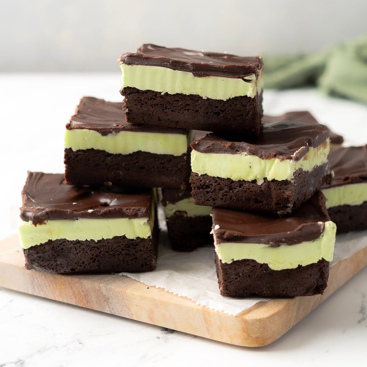 Several layered mint chocolate brownies with a green mint filling and chocolate topping are stacked on a wooden board lined with parchment paper.