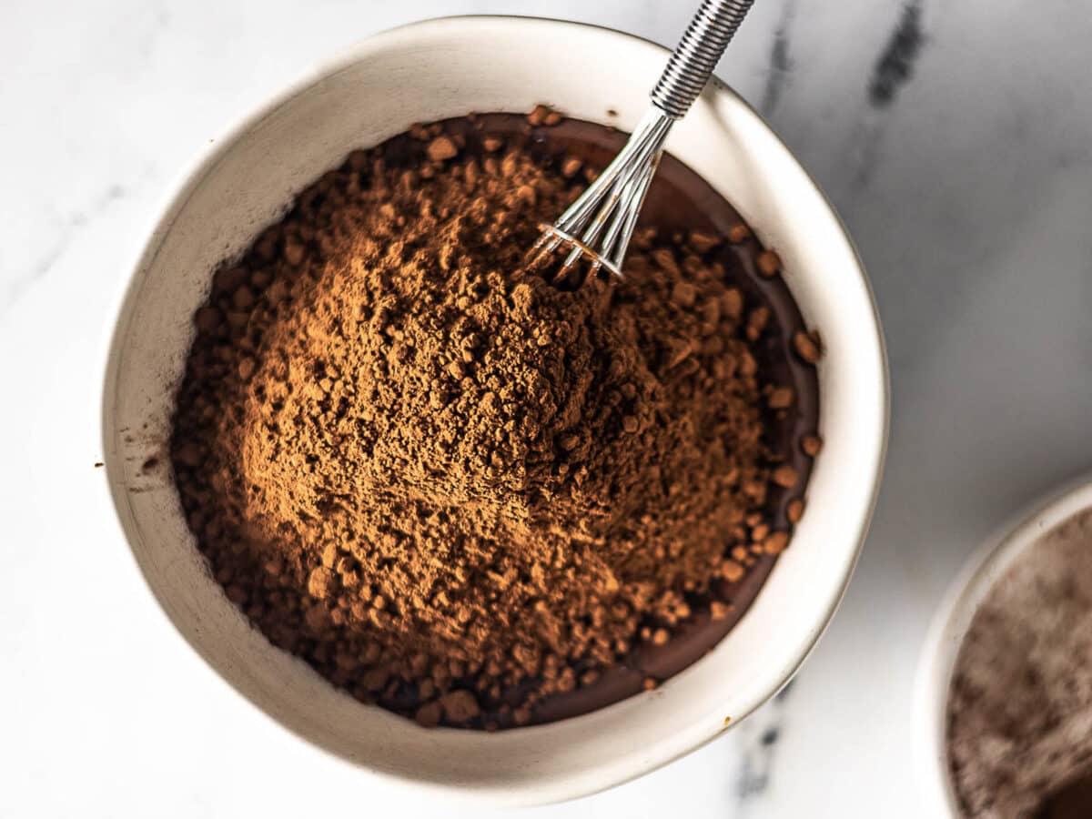 A bowl filled with cocoa powder on top of melted chocolate, with a small metal whisk resting inside.