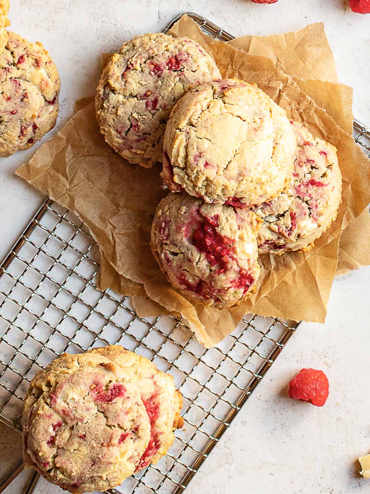 Several raspberry cookies are arranged on parchment paper atop a cooling rack, with a few cookies and raspberries scattered nearby on a white surface.