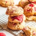 Three raspberry scones are stacked on a cooling rack, with more scones and a single raspberry nearby.