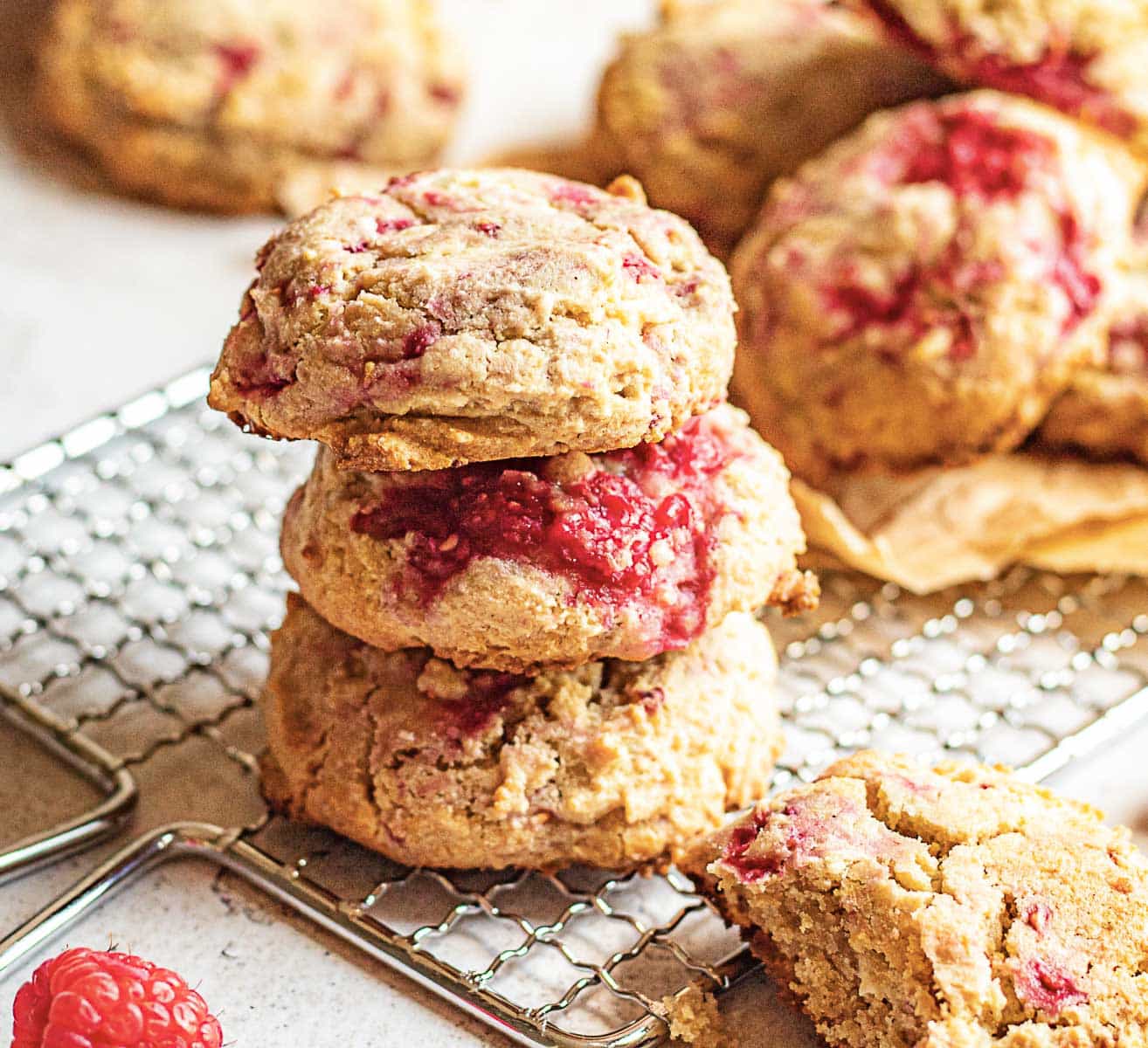 Three raspberry scones are stacked on a cooling rack, with more scones and a single raspberry nearby.