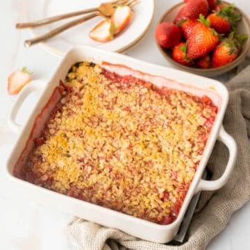 A baked strawberry crumble in a white square dish, with a bowl of fresh strawberries and a plate with forks and sliced strawberries beside it.