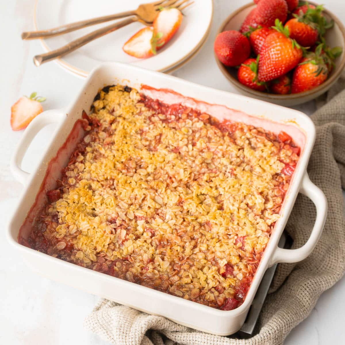 A baked strawberry crumble in a white square dish, with a bowl of fresh strawberries and a plate with forks and sliced strawberries beside it.