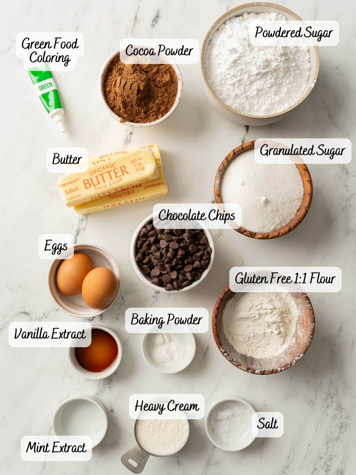 Various labeled baking ingredients, including eggs, butter, sugars, cocoa powder, chocolate chips, flour, green food coloring, extracts, baking powder, heavy cream, and salt on a white surface.