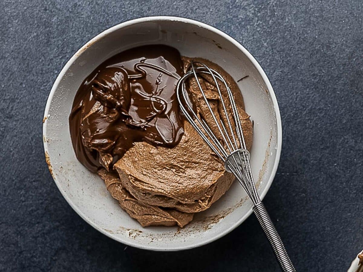 A bowl with chocolate mousse mixture and melted chocolate, with a metal whisk resting inside, on a dark countertop.