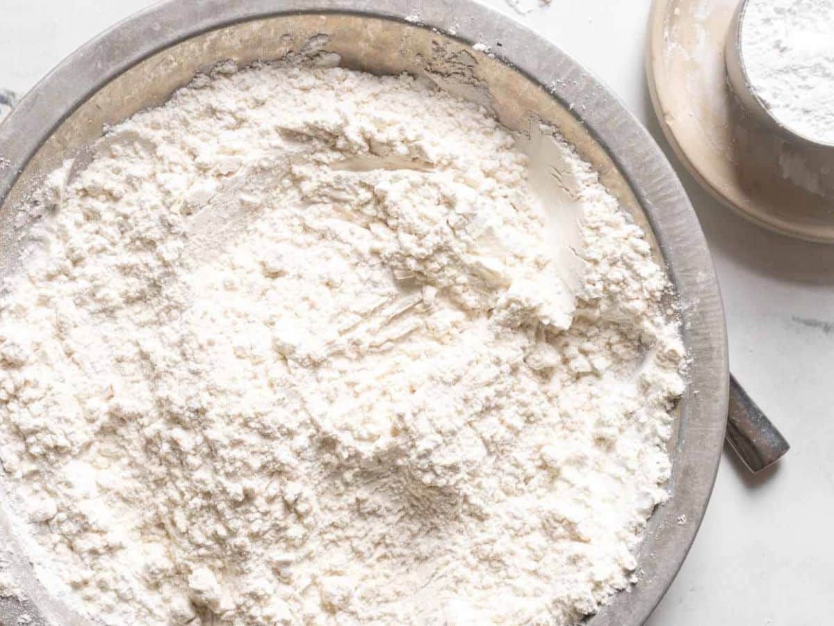 A metal bowl filled with white flour, with a smaller bowl of flour beside it on a white surface.