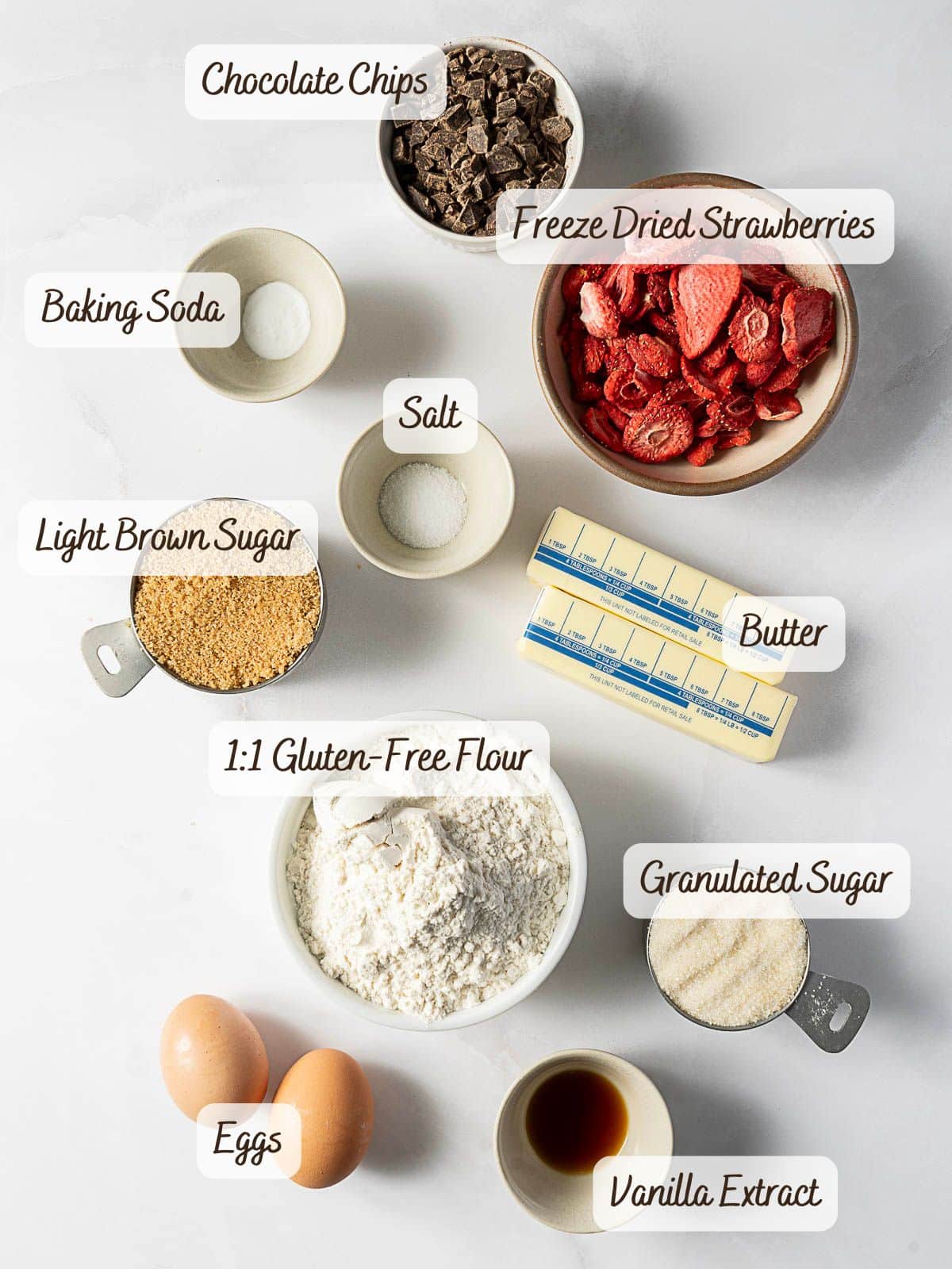 Ingredients for baking gluten free strawberry cookies are arranged on a white surface, including chocolate chips, freeze dried strawberries, butter, sugar, brown sugar, gluten free flour, eggs, vanilla extract, baking soda, and salt.
