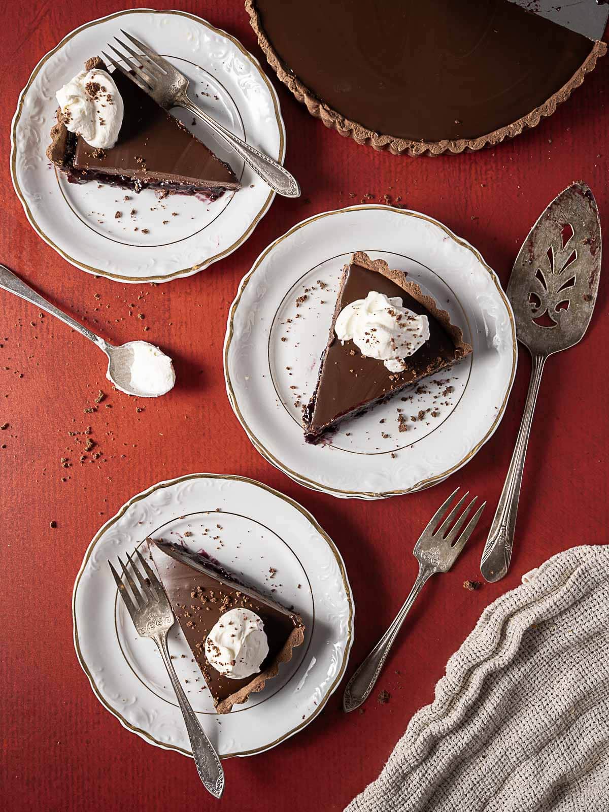 Three slices of chocolate tart with whipped cream on white plates, served with forks and dessert spoons on a red surface.