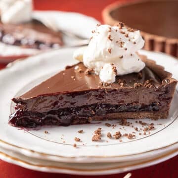 A slice of chocolate and cherry pie topped with whipped cream sits on a white plate, with another slice and the pie in the background.