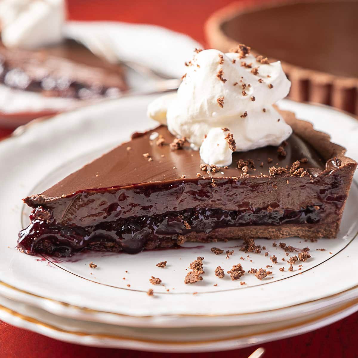 A slice of chocolate and cherry pie topped with whipped cream sits on a white plate, with another slice and the pie in the background.