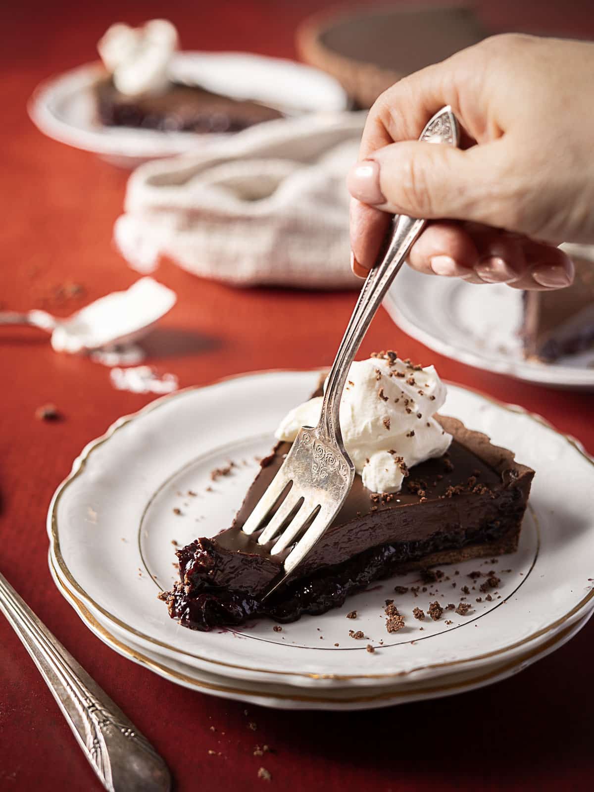 A hand holds a fork, taking a bite from a slice of chocolate pie topped with whipped cream on a white plate.