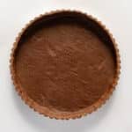 Unbaked chocolate tart crust with fork marks in a fluted tart pan, viewed from above on a white surface.
