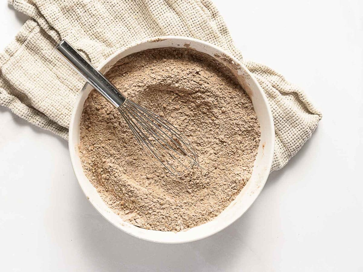 A metal whisk rests in a white bowl filled with blended dry cocoa powder mix, placed on a beige textured cloth against a white background.