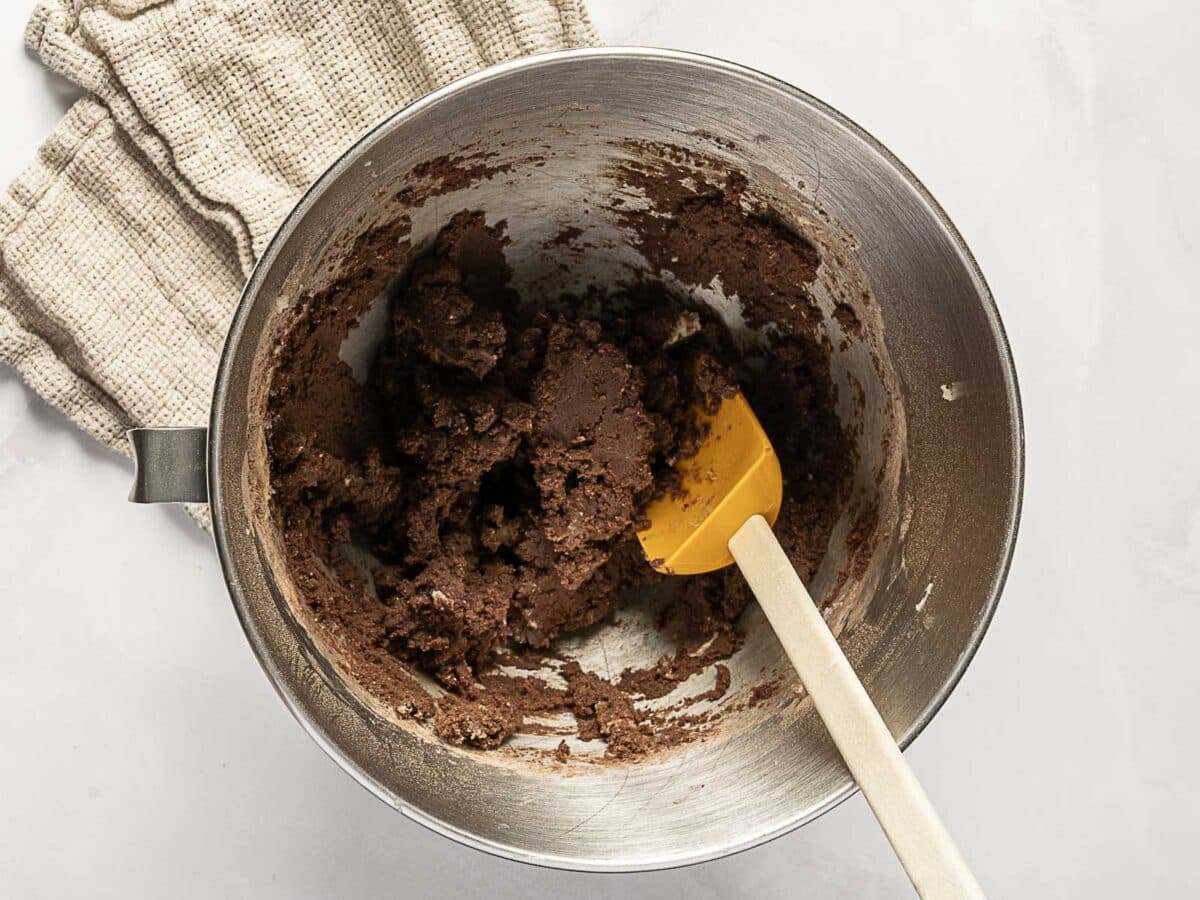 A mixing bowl containing chocolate cookie dough with a yellow spatula, placed on a light surface next to a folded beige kitchen towel.