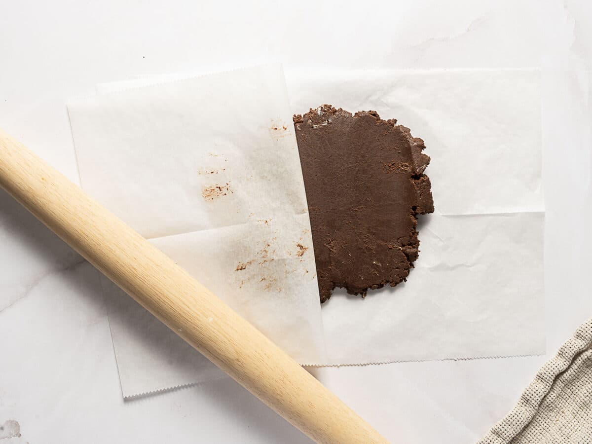 A rolling pin beside chocolate dough flattened between two sheets of parchment paper on a light-colored surface.